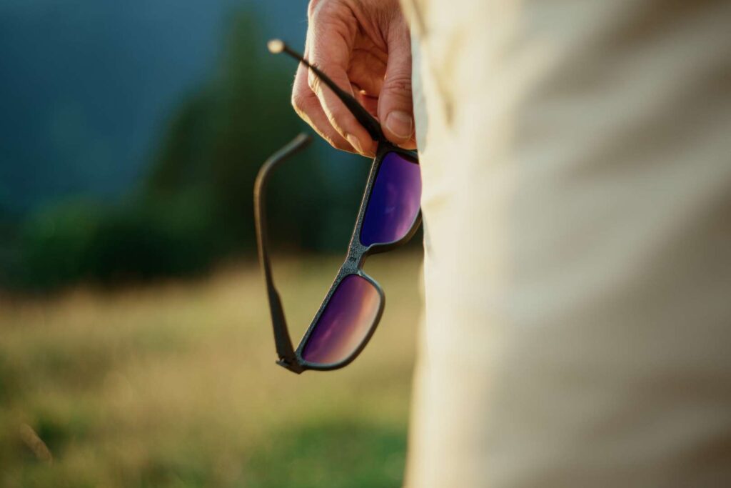Close-up of a hand holding funky sunglasses with purple lenses against a blurred natural background.
