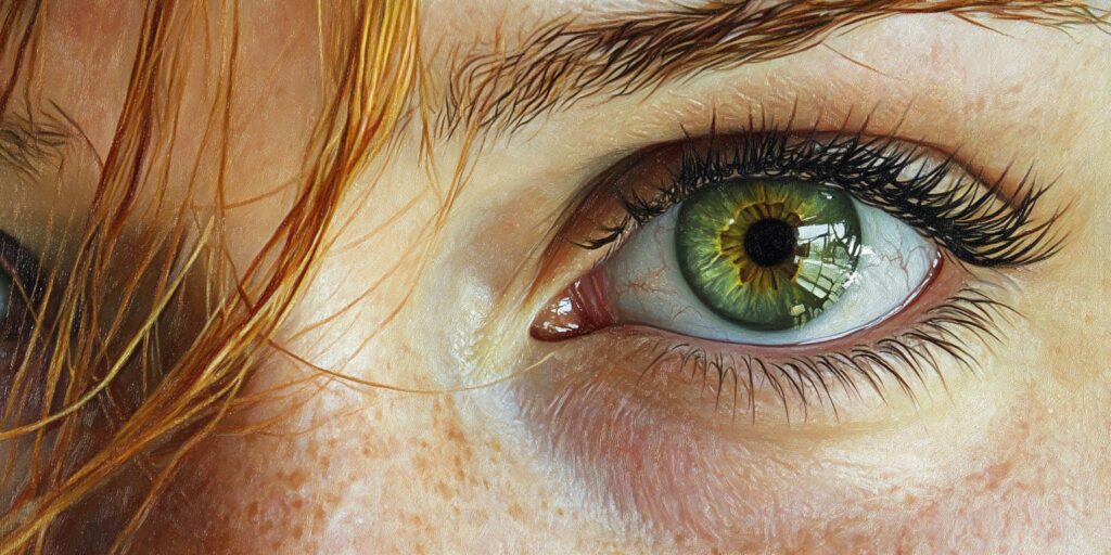 Close-up of a green eye with a bright yellow center, detailed eyelashes, and subtle reflections. The eye is framed by natural freckles and strands of red hair.