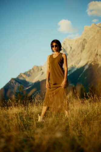 Frau mit Sonnenbrille steht in Berglandschaft auf einer Wiese im goldenen Abendlicht